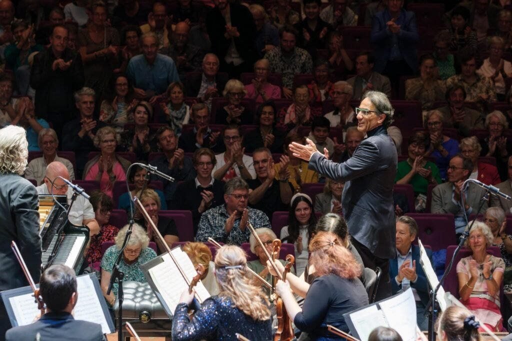 Alvaro Siviero de pé diante da orquestra, aplaudindo os músicos, com a plateia também de pé